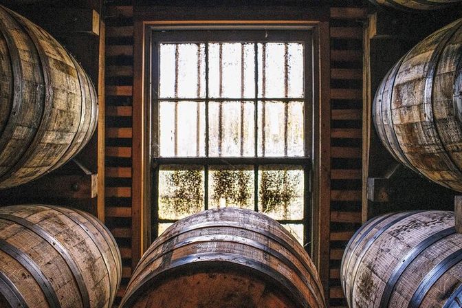 Oak whiskey barrels stacked in a dim distillery aging room around a weathered window with sunlight filtering through.