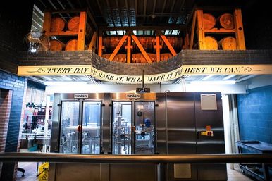 Craft brewery interior showing stainless-steel bottling line beneath stacked wooden barrels on a mezzanine and a curved slogan banner