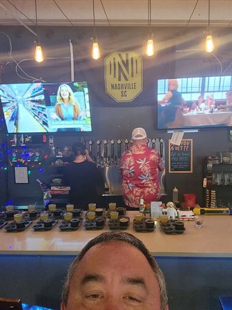 Selfie view of a lively Nashville bar interior with beer flight samples lined on the counter, a row of draft taps, a patron in a red Hawaiian shirt, two TV screens and a Nashville SC logo under warm hanging bulbs.