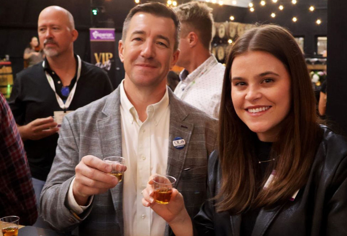 Smiling man and woman holding small whiskey sample cups at an indoor tasting event with string lights and a casual networking crowd in the background.