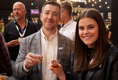 Smiling man and woman holding small whiskey sample cups at an indoor tasting event with string lights and a casual networking crowd in the background.
