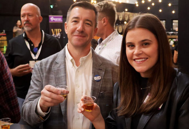 Smiling man and woman holding small whiskey sample cups at an indoor tasting event with string lights and a casual networking crowd in the background.