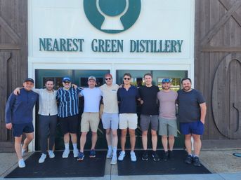 Group of nine men posing arm-in-arm outside a distillery entrance with a large green logo overhead, wearing casual summer clothes, shorts and sunglasses in a daytime group photo.