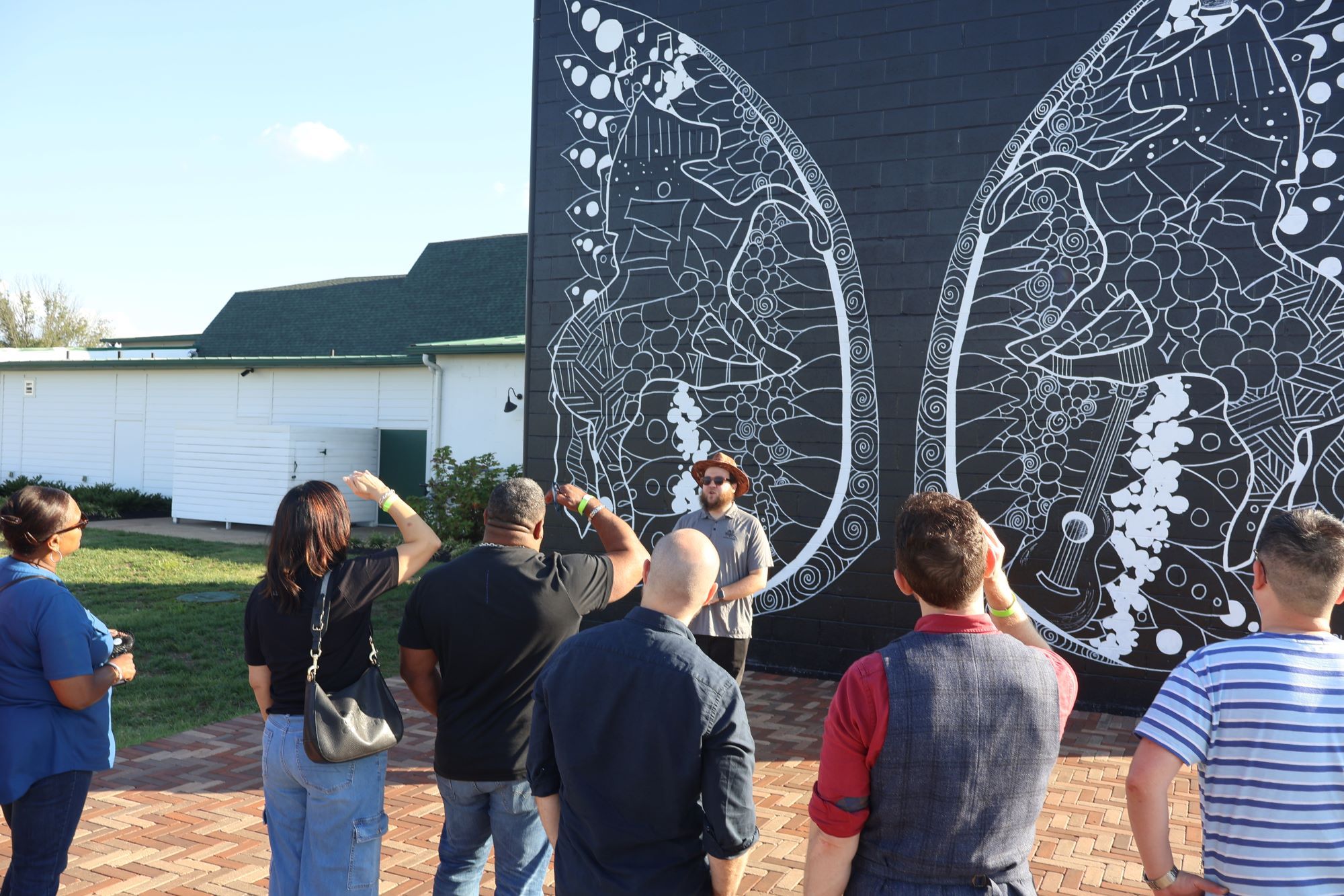 Group on a sunny outdoor art tour in a public plaza admiring a large black-brick street-art mural of intricate white butterfly wings
