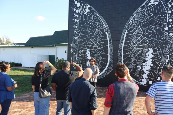 Group on a sunny outdoor art tour in a public plaza admiring a large black-brick street-art mural of intricate white butterfly wings