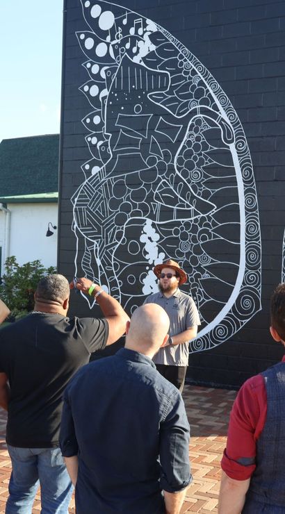 Group on a sunny outdoor art tour in a public plaza admiring a large black-brick street-art mural of intricate white butterfly wings