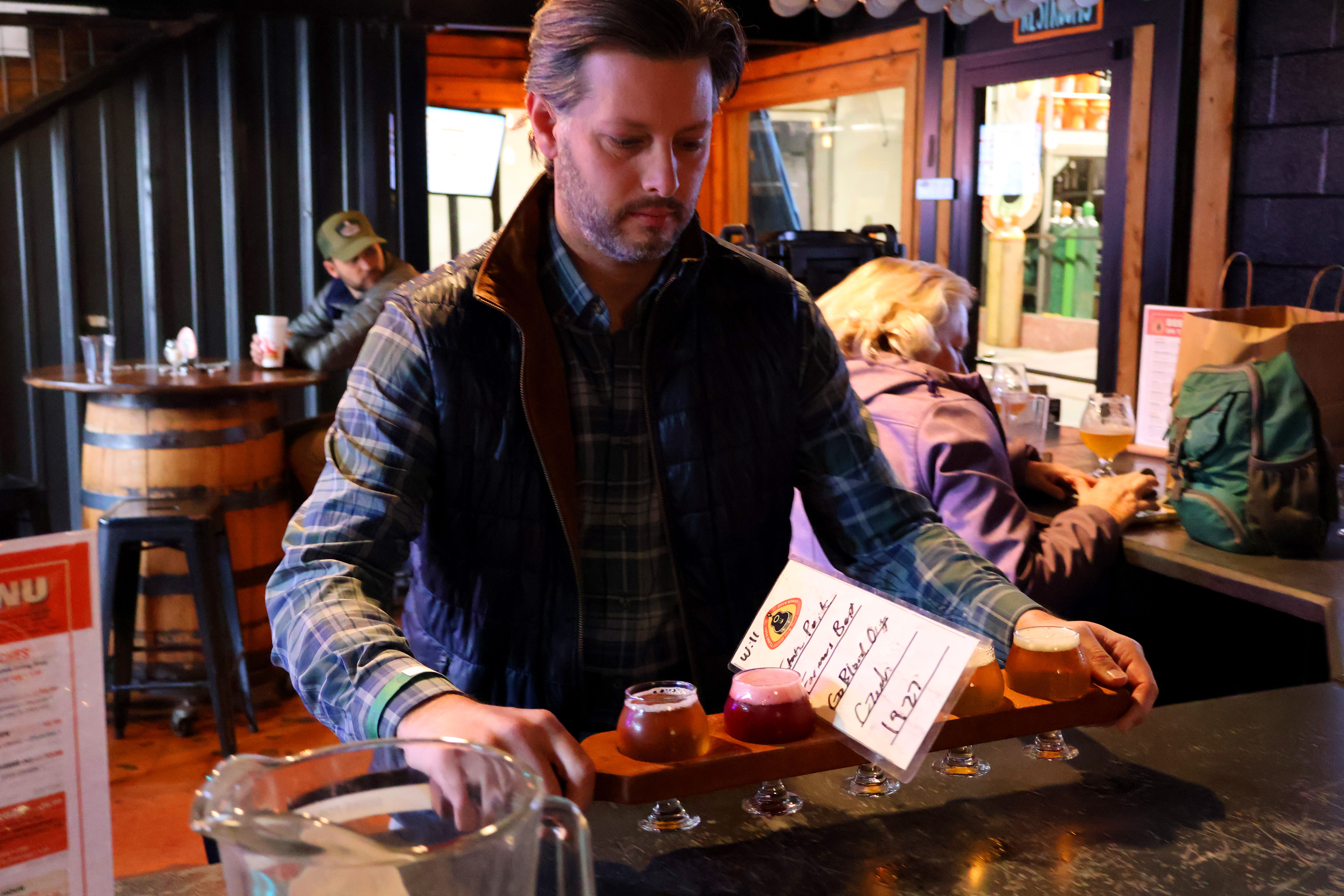 Person carrying a wooden flight of colorful craft beer tasters across a cozy brewery taproom bar, ready for a tasting.