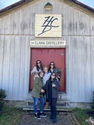 Five friends laughing and holding drinks outside a rustic wooden distillery barn with a red door and large logo sign.