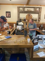 Rustic distillery tasting room with wooden bar and shelves of spirits, staff serving amber liquor, a tasting flight of small plastic cups on the counter and patrons at the bar