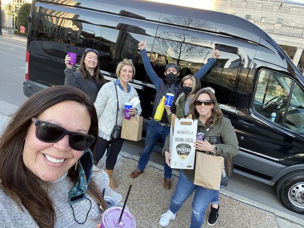 Six friends taking a daytime selfie in front of a black passenger van on a downtown street, holding drinks, paper takeout bags and a six-pack carrier, smiling and cheering.