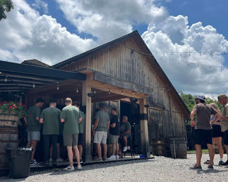 Group of people in casual summer clothes gathered at a rustic barn-style outdoor bar with string lights, wooden barrels, flower planters, gravel courtyard and dramatic cloudy blue sky.