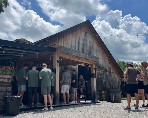Group of people in casual summer clothes gathered at a rustic barn-style outdoor bar with string lights, wooden barrels, flower planters, gravel courtyard and dramatic cloudy blue sky.