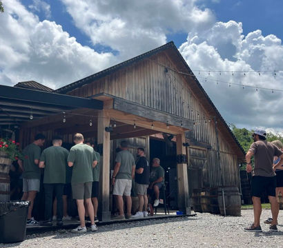 Group of people in casual summer clothes gathered at a rustic barn-style outdoor bar with string lights, wooden barrels, flower planters, gravel courtyard and dramatic cloudy blue sky.