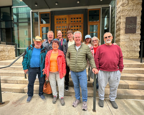 Smiling group of nine adults in colorful jackets posing on stone steps outside a downtown building with wooden doors and glass facade.