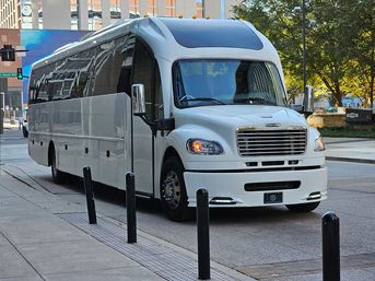 Sleek white charter shuttle bus parked on a downtown city street, front three-quarter view beside a sidewalk with black bollards and modern buildings and trees in the background.