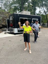 Cheerful woman in a lime-green polo holding a cold drink and smiling in front of a black food truck at a tree-lined park, with another person behind her also holding a beverage.