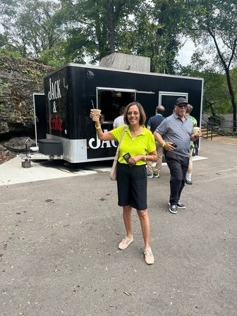 Cheerful woman in a lime-green polo holding a cold drink and smiling in front of a black food truck at a tree-lined park, with another person behind her also holding a beverage.