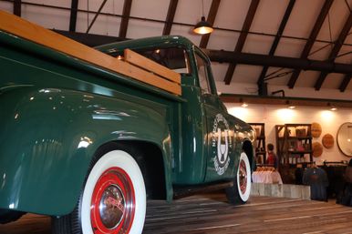 Vintage green pickup truck with wooden bed rails and red-rimmed whitewall tires displayed on a wooden platform inside a rustic retail space with exposed beams and shoppers browsing clothing.