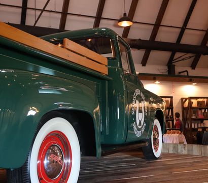Vintage green pickup truck with wooden bed rails and red-rimmed whitewall tires displayed on a wooden platform inside a rustic retail space with exposed beams and shoppers browsing clothing.