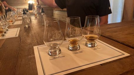Three whiskey tasting glasses with amber pours on a labeled flight mat atop a rustic wooden bar, ready for tasting in a cozy distillery tasting room.