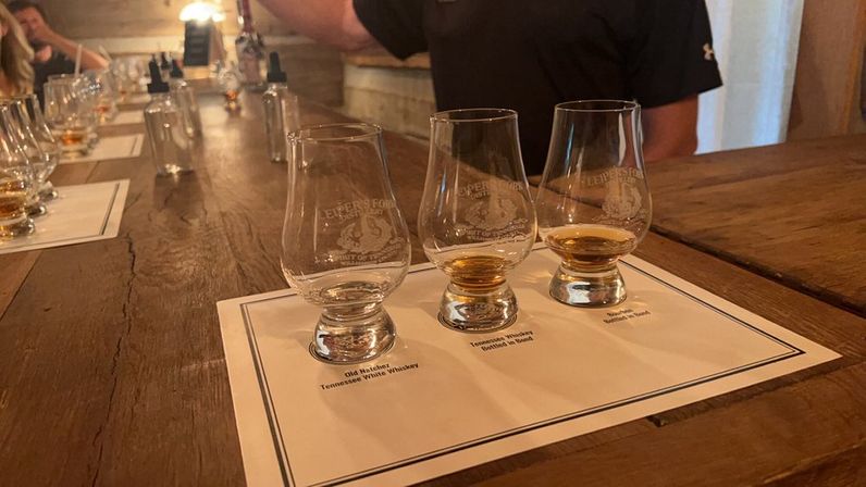 Three whiskey tasting glasses with amber pours on a labeled flight mat atop a rustic wooden bar, ready for tasting in a cozy distillery tasting room.