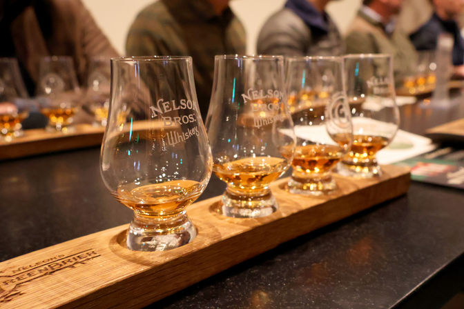 Whiskey tasting flight of four Glencairn-style glasses with amber whiskey on a wooden paddle in a distillery tasting room, blurred patrons at the bar in the background.