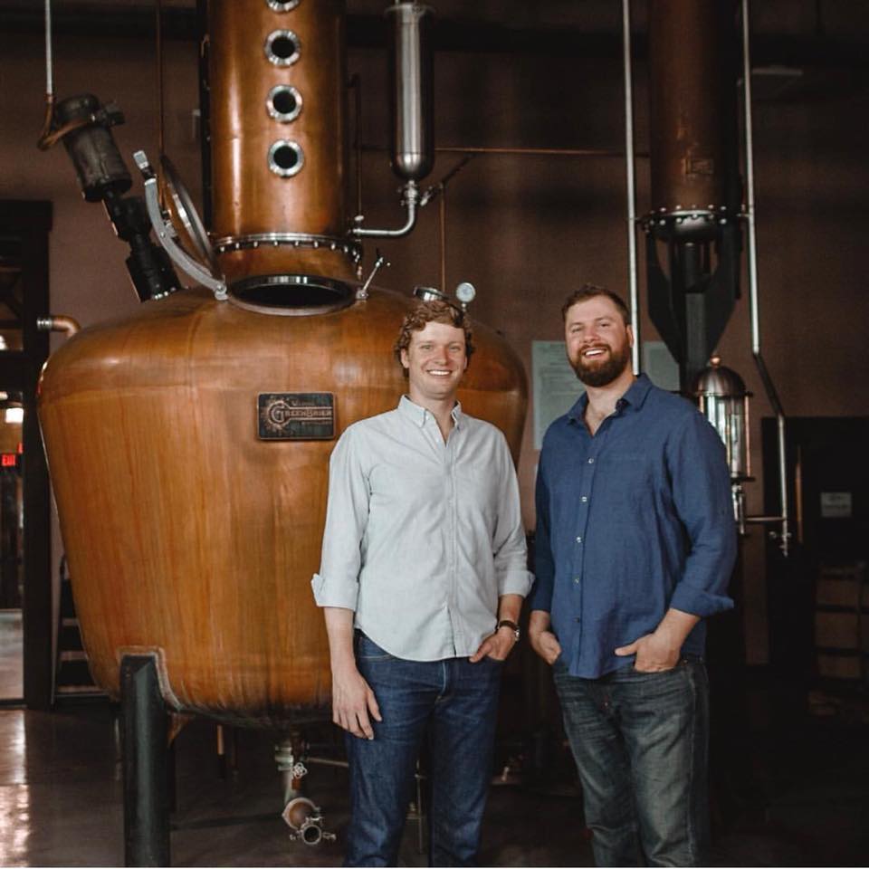 Two smiling men in casual shirts pose beside a large copper pot still in a craft distillery production room, evoking small-batch spirits making.