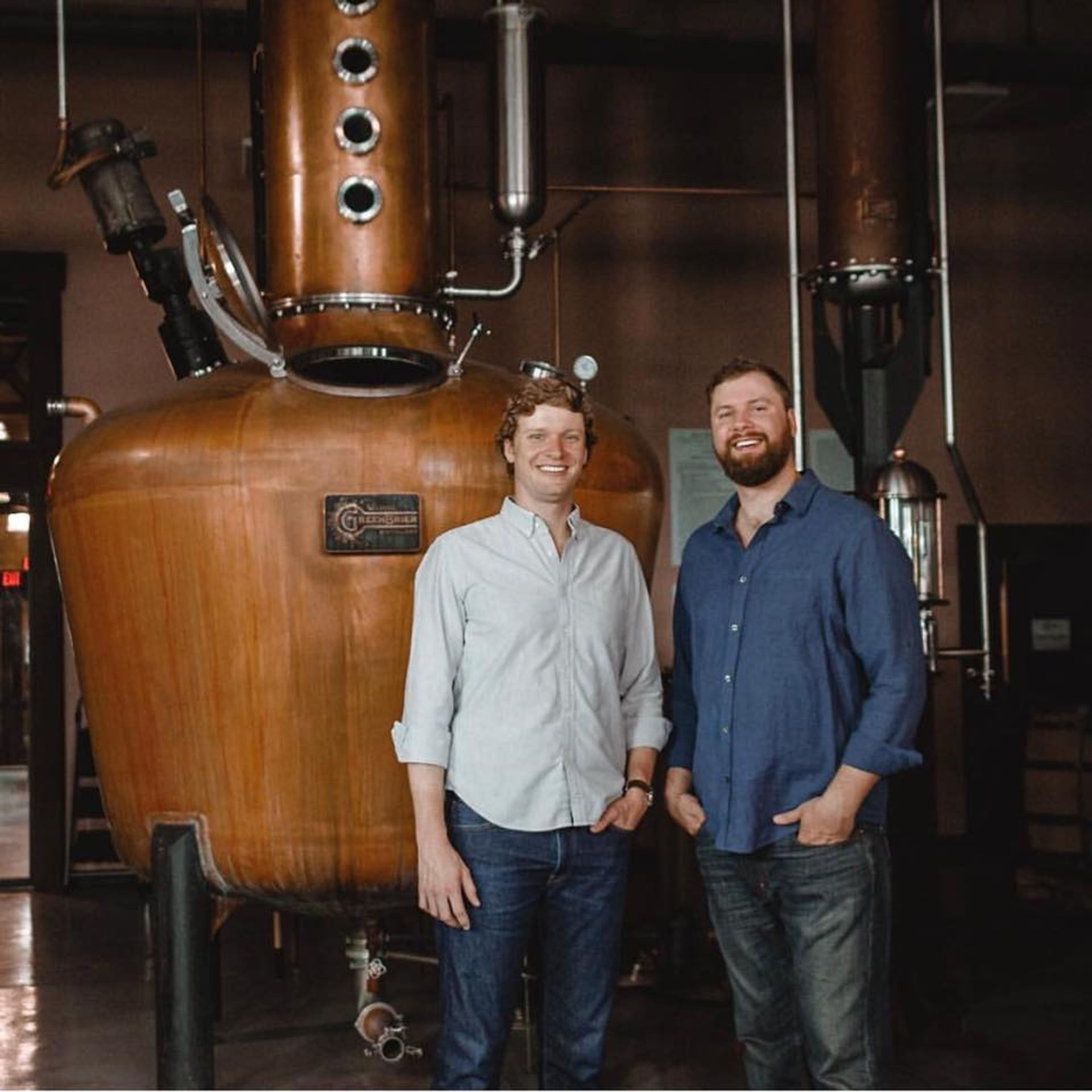 Two smiling men in casual shirts pose beside a large copper pot still in a craft distillery production room, evoking small-batch spirits making.