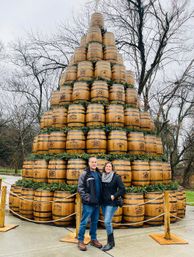 Two smiling visitors posing in front of a towering outdoor pyramid of stacked wooden barrels decorated with evergreen garlands and rope barriers, set against leafless trees on an overcast winter day.