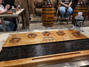Whiskey tasting flight on a wooden paddle with four logoed sample glasses, bottled water and oak barrels in a distillery tasting room.