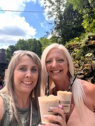 Close-up selfie of two smiling women holding frozen summer cocktails outdoors on a sunny day at a wooded patio with a rocky wall and green trees