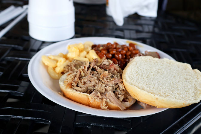 Open pulled-pork sandwich with shredded pork, macaroni and cheese and baked beans on a white disposable plate sitting on a black outdoor table — casual BBQ plate.