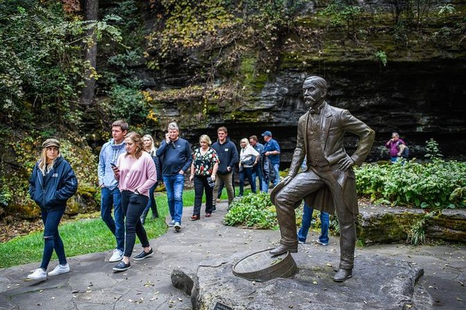 Bronze statue of a 19th-century gentleman with one foot on a barrel on a rocky outcrop, beside a paved scenic gorge trail where visitors stroll among limestone cliffs and green foliage.