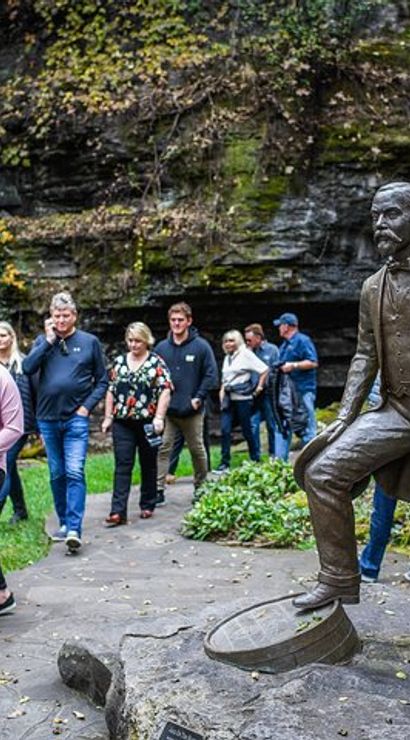 Bronze statue of a 19th-century gentleman with one foot on a barrel on a rocky outcrop, beside a paved scenic gorge trail where visitors stroll among limestone cliffs and green foliage.