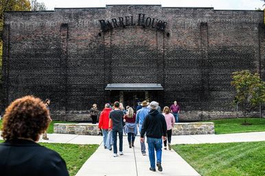 Group of visitors walking along a paved path toward the entrance of a large historic brick warehouse with a metal sign above the door, surrounded by green lawn and trees