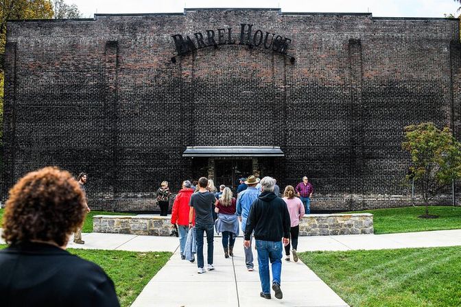 Group of visitors walking along a paved path toward the entrance of a large historic brick warehouse with a metal sign above the door, surrounded by green lawn and trees