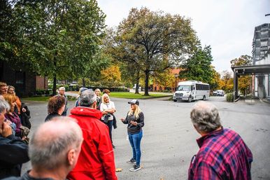 Tour guide addressing a group of adults outdoors on a crisp autumn day, tree-lined campus street with fall foliage and a white shuttle bus parked nearby