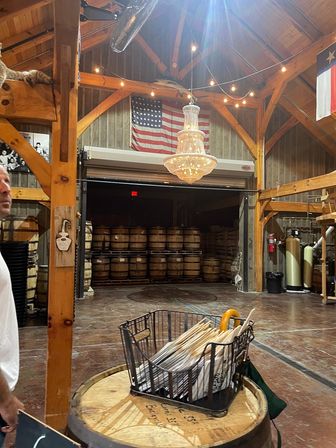 Rustic barn-style tasting room/event space with exposed timber beams, warm string lights, a sparkling crystal chandelier, stacked oak barrels, an American flag, and a basket of menus and umbrellas on a barrel.