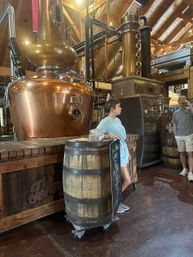Craft distillery tasting room with oversized copper pot stills and column, wood‑beamed ceiling, wooden barrels used as display tables and a chalkboard menu, with casually dressed visitors nearby.