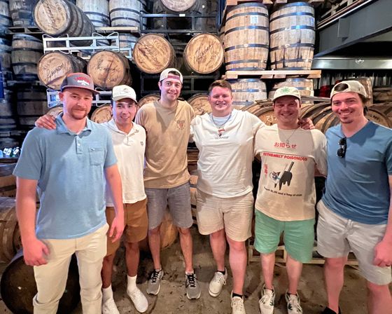 Six men smiling and posing in a distillery barrel room in front of stacked wooden bourbon barrels and racks, wearing casual summer clothes and caps — group tour photo.