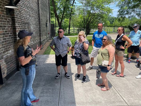 Animated tour guide speaking to a small group on a sunlit concrete patio beside a historic brick building, casual summer attire with trees and parkland visible in the background.