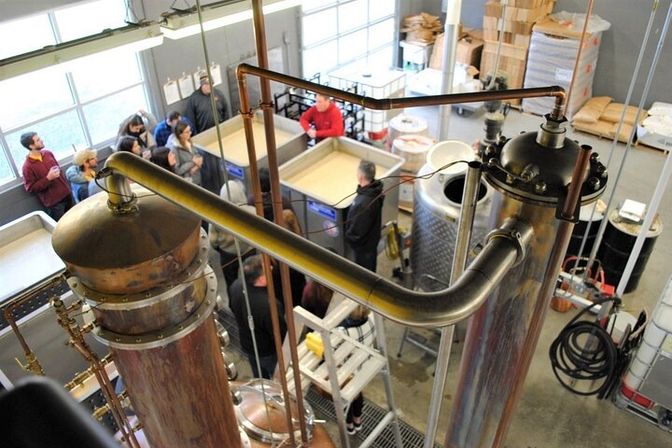 Overhead view of a craft distillery interior showing large copper pot stills, stainless fermentation tanks and a group of visitors on a guided tour