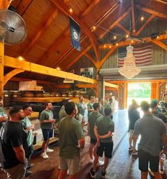 Casual group gathered inside a sunlit rustic barn-style event space with exposed timber beams, a hanging crystal chandelier, string lights, and an American flag over the open doorway.