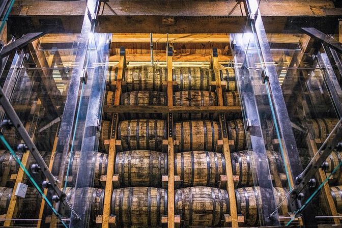 Stacked oak whiskey barrels on wooden racks behind glass in a distillery warehouse aging room, rustic industrial vibe
