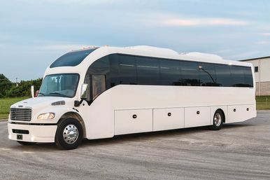 Sleek white charter coach bus with tinted windows parked on a paved lot by an industrial building under a cloudy sky