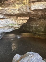 Layered limestone cave interior with a small cascading waterfall flowing into a dark natural pool and a foreground boulder
