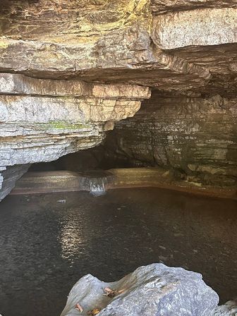 Layered limestone cave interior with a small cascading waterfall flowing into a dark natural pool and a foreground boulder