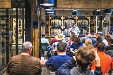 Group at a rustic distillery whiskey tasting, guide holding up a bottle in front of stacked oak barrels in a barrel room