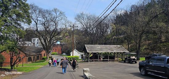 People walking down a paved lane toward red-brick buildings and a rustic open wooden pavilion beside parked vehicles, set against a wooded hillside and blue sky.