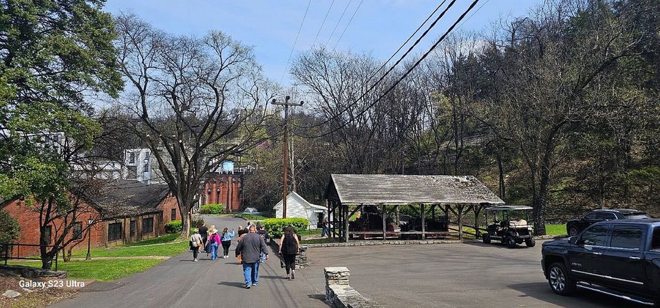People walking down a paved lane toward red-brick buildings and a rustic open wooden pavilion beside parked vehicles, set against a wooded hillside and blue sky.